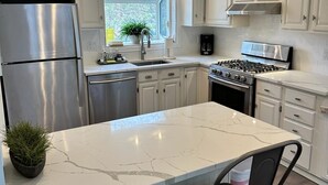 Cozinha moderna com armários brancos, eletrodomésticos de aço inoxidável, bancada de mármore e janela que permite entrada de luz natural.