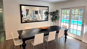 Uma área de refeições com mesa preta, cadeiras brancas, um espelho grande e vista para uma piscina através de portas de vidro.