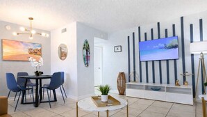 A modern living room with a dining area, a flat-screen TV mounted on the wall, and a coffee table with a plant. - Family and pet friendly ~ Pool, gym, games! (Indian Harbour Beach)