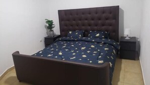 Ein Schlafzimmer mit einem dunkelbraunen, gepolsterten Kopfteil, einem Bett mit einer blauen Bettdecke mit Blattmuster, zwei Nachttischen mit Lampen und einer Zimmerpflanze auf einem Tisch.