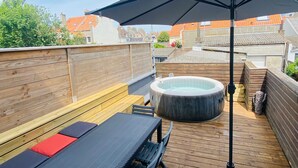 Een houten terras met een jacuzzi, een tafel en banken.