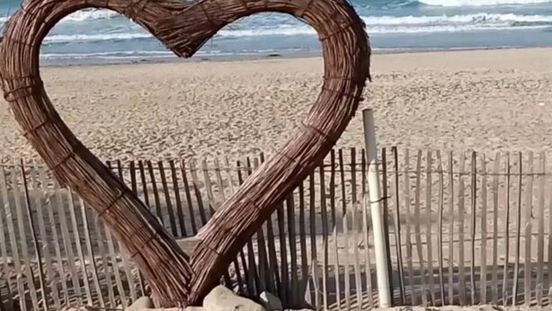 Een hartvormige constructie van takken op een zandstrand, met een houten hek op de achtergrond en de oceaan in de verte.