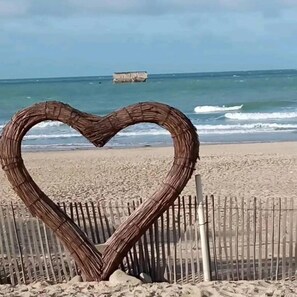 A heart-shaped structure made of sticks on a sandy beach with a wooden fence in the background and the ocean in the distance.