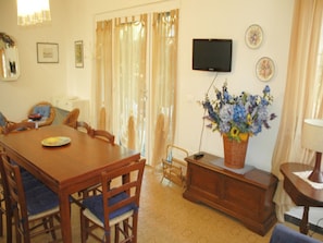 A dining area with a wooden table and chairs, a television mounted on the wall, and a floral arrangement on a side table.