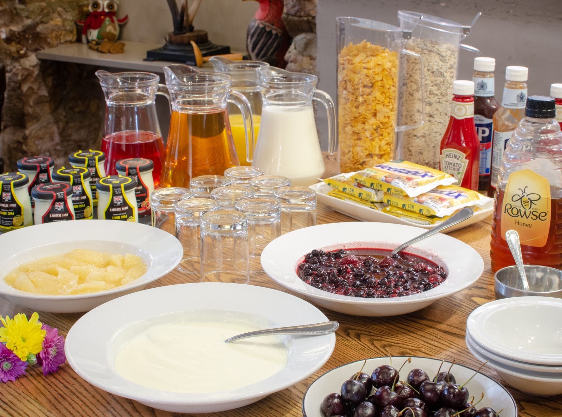 A table with various food items and condiments, including bowls of yogurt, a bowl of cherries, and a pitcher of honey.