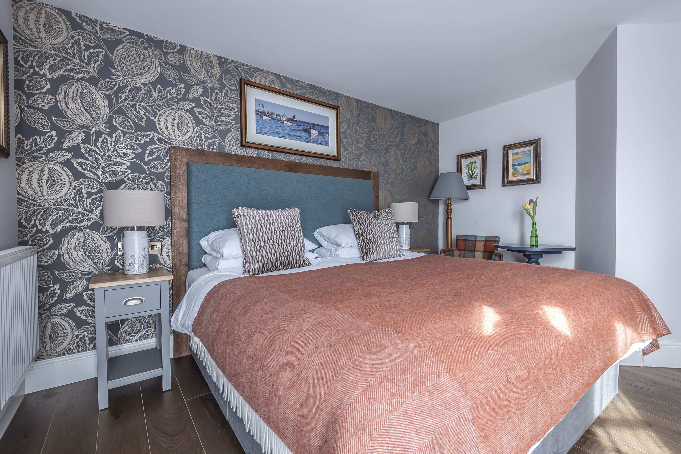 A neatly arranged bedroom with a large bed, a bedside table with a lamp, a small table with a vase, and a framed picture on the wall.