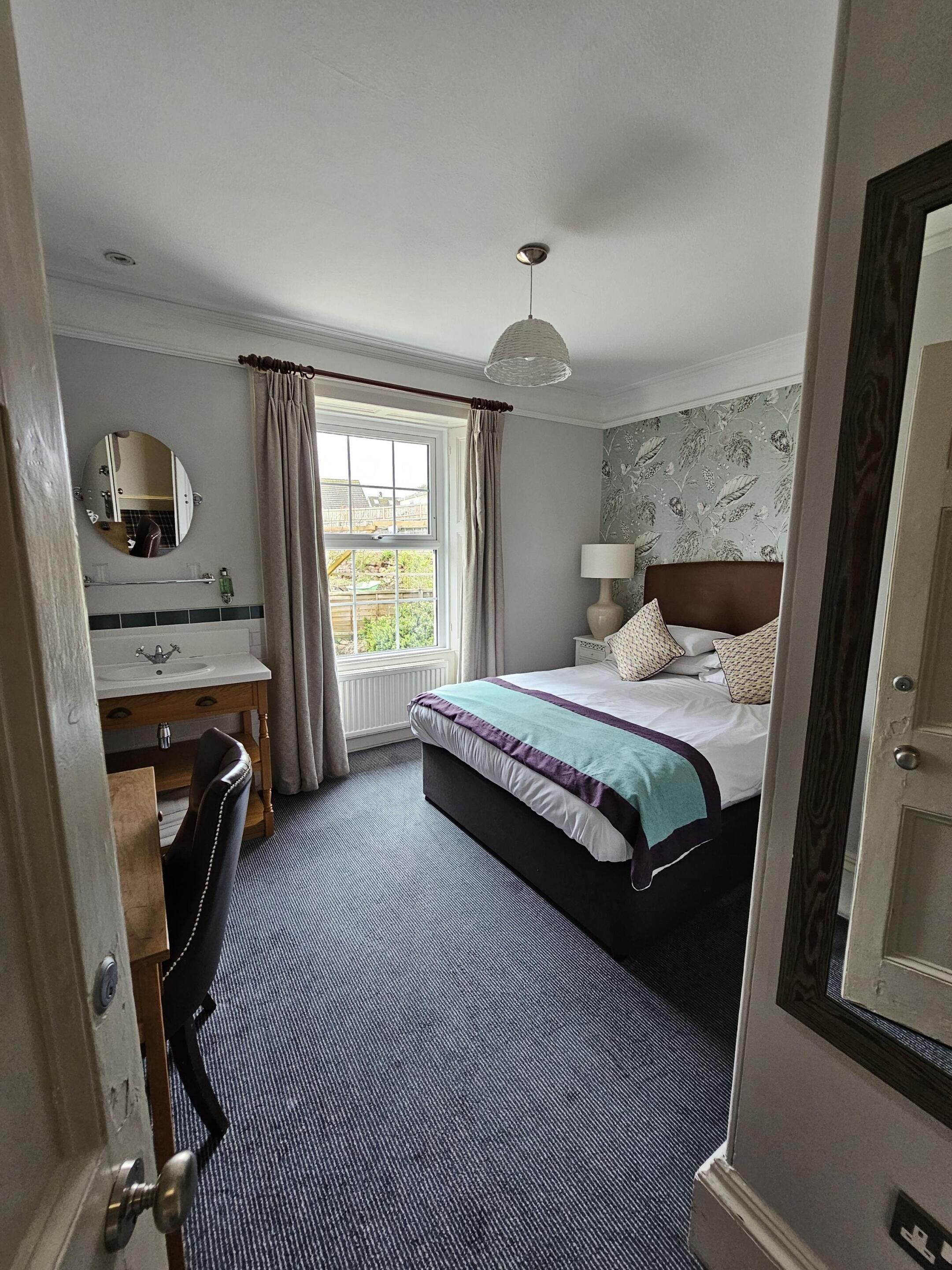 A hotel room with a bed, bedside table, lamp, and a view of the outside.