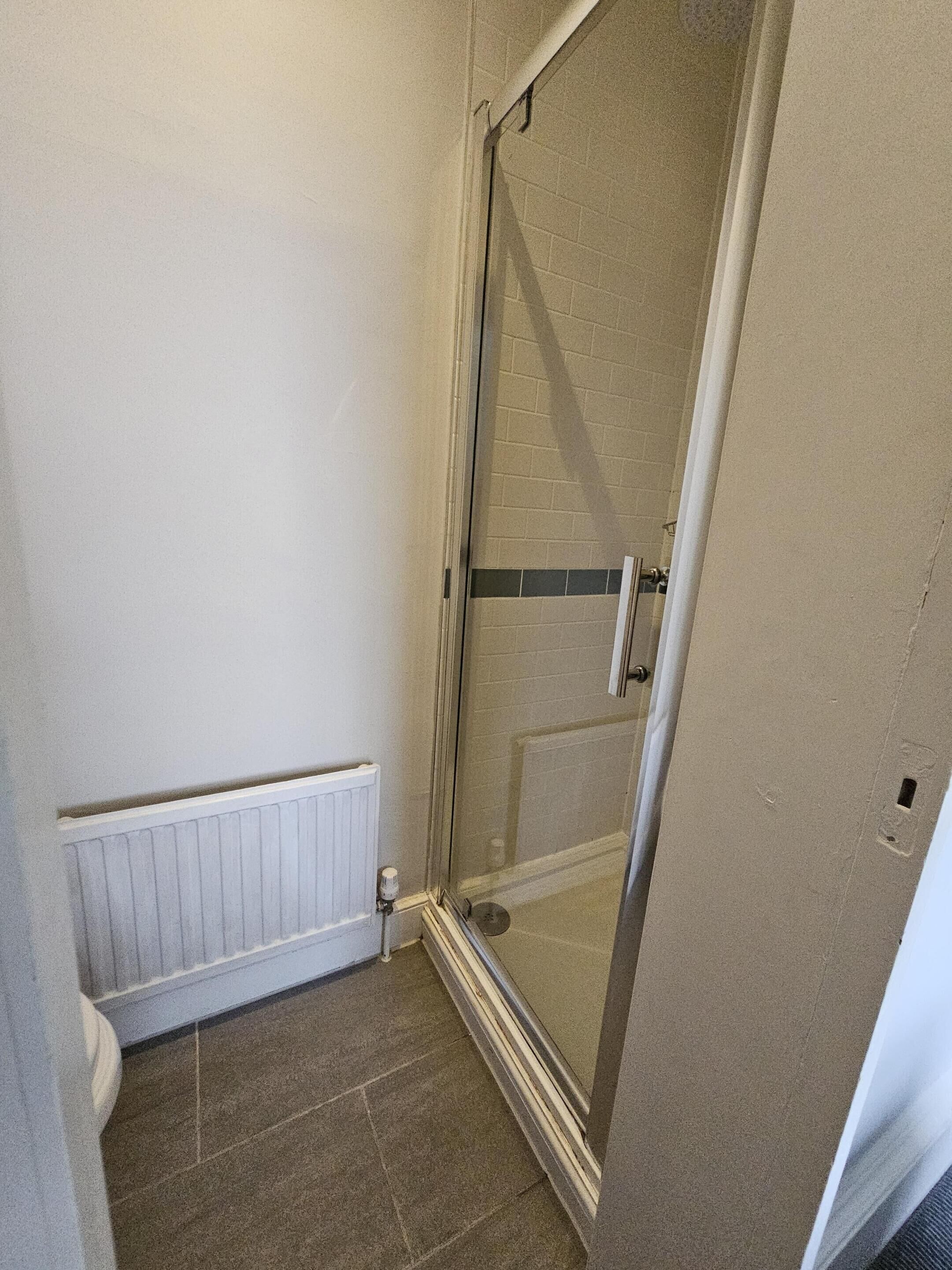 Un espacio de ducha pequeño y cerrado con puerta de vidrio, un radiador blanco y suelo de baldosas.