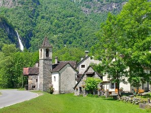 A stone church with a bell tower, surrounded by lush greenery and a waterfall in the background.