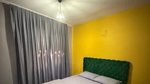 Una cama bien tendida con un cabecero verde, una toalla enrollada y una ventana con cortinas grises.
