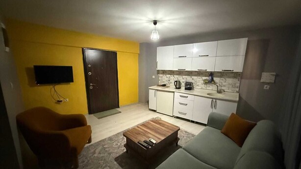 Un espacio habitable compacto con sofá, dos sillones, una mesita de centro, televisor, una zona de cocina con armarios y una puerta.