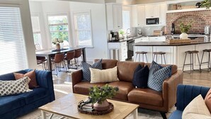A spacious living room with a brown leather sofa, blue armchairs, and a coffee table. A kitchen with white cabinets and a brick backsplash is visible in the background. - Luxury - 6000sqft- Sleeps 22+ Basketball, Billiards, Theater, Game, PrincessRoom (Layton)