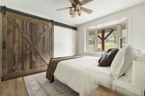 A bedroom with a large wooden barn door, a ceiling fan, a bed with white bedding, and a view of the outdoors through a window. - Amazing views, hot tub/outdoor fireplace, countryside, farm with calves & goats. (Navarre)
