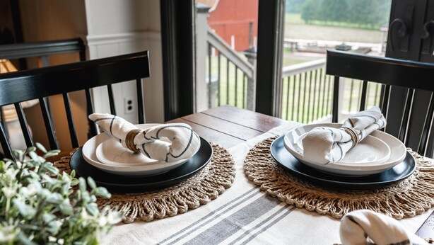 A dining table with two plates and napkins, set on a woven placemat, with a view of a red barn and greenery outside. - Amazing views, hot tub/outdoor fireplace, countryside, farm (Navarre)