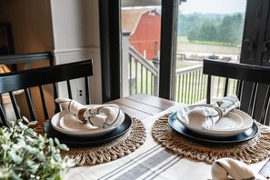 A dining table with two plates and napkins, set on a woven placemat, with a view of a red barn and greenery outside. - Amazing views, hot tub/outdoor fireplace, countryside, farm with calves & goats. (Navarre)
