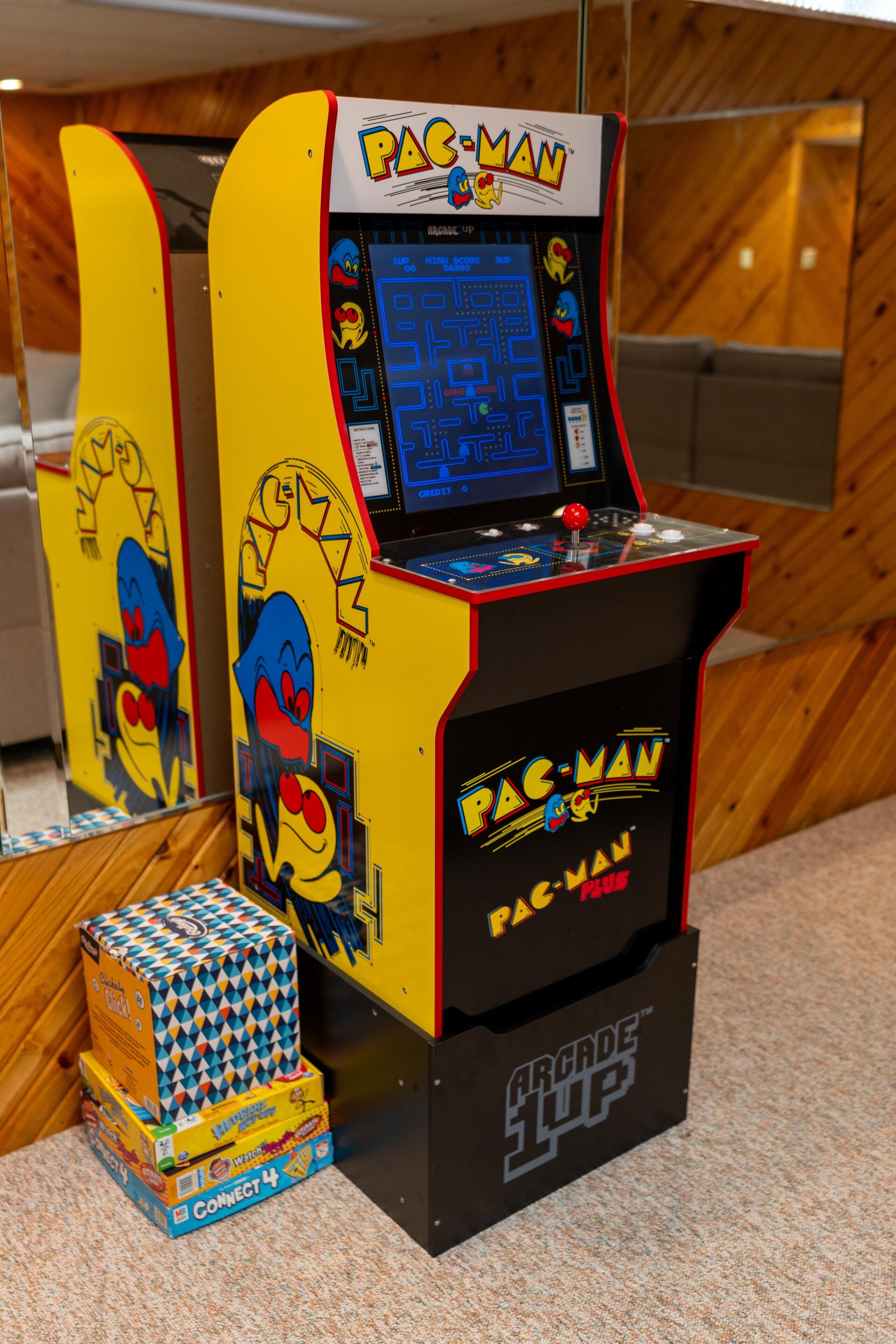 Deux bornes d’arcade Pac-Man aux designs symétriques, une petite pile de boîtes de jeux et un mur en bois en arrière-plan.