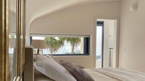 Une chambre à coucher avec un lit, des oreillers, une table de chevet agrémentée d’objets décoratifs et une vue sur des palmiers par la fenêtre.