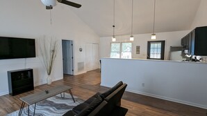 A spacious living area with a wooden floor, a flat-screen TV, a coffee table, and a sofa. There is a kitchen area with a refrigerator, cabinets, and a window.
