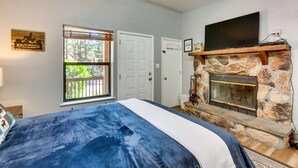 A bedroom with a bed, a stone fireplace, a TV mounted above it, and a window with blinds. - Angel Fire Studio w/ Fireplace, Walk to Resort! (Angel Fire)