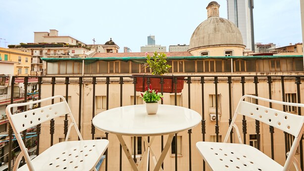 Un balcón con dos sillas blancas y una mesita, una planta en maceta y vistas de edificios de la ciudad.