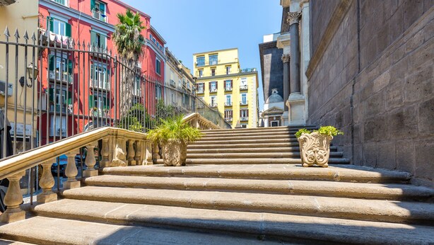Un escalier en pierre menant à un bâtiment agrémenté d’une rampe en fer forgé et de jardinières décoratives.