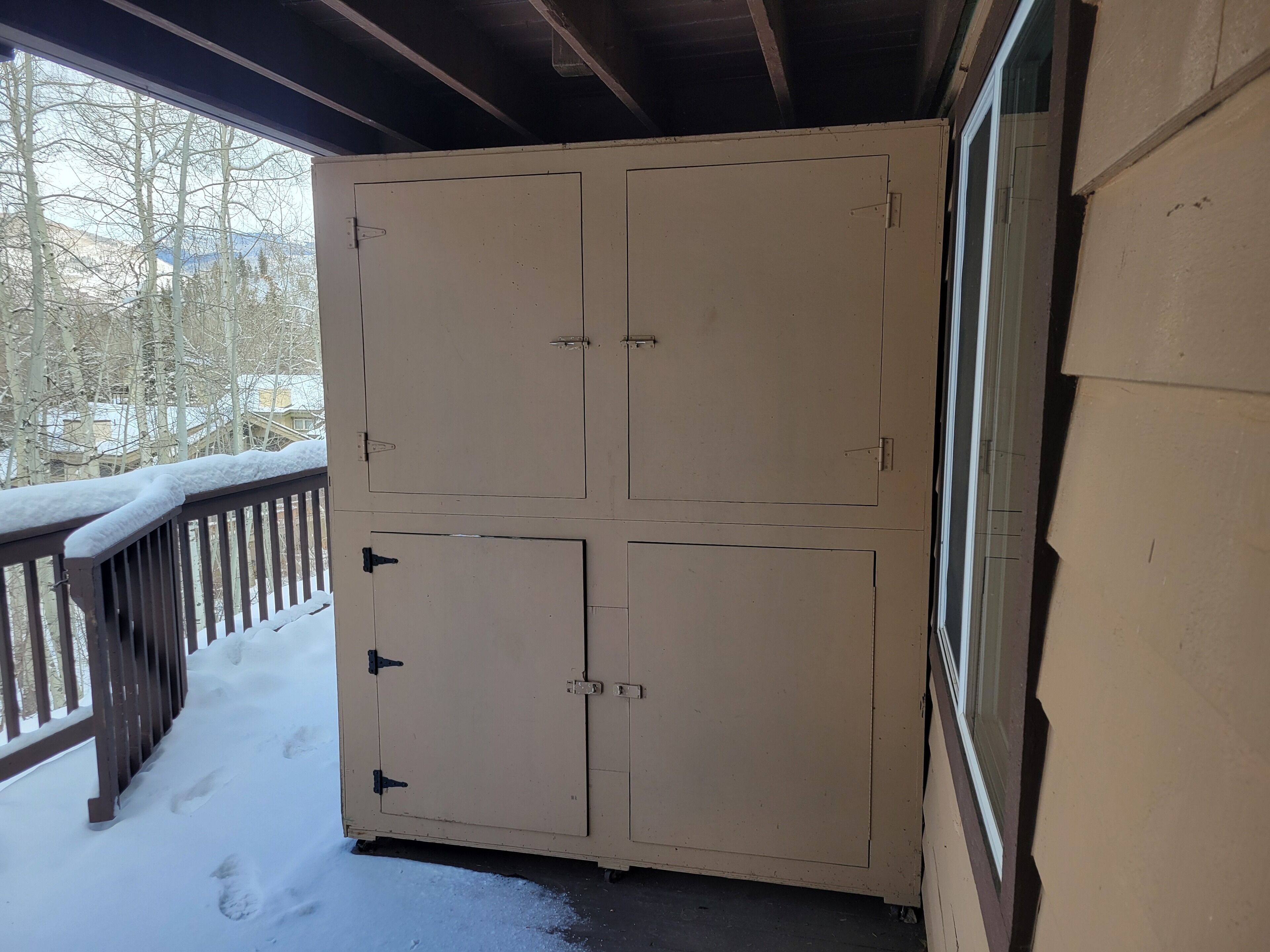 Ett stort, beigefärgat förvaringsskåp i metall med fyra dörrar, placerat på en balkong med ett träräcke och snö på marken.