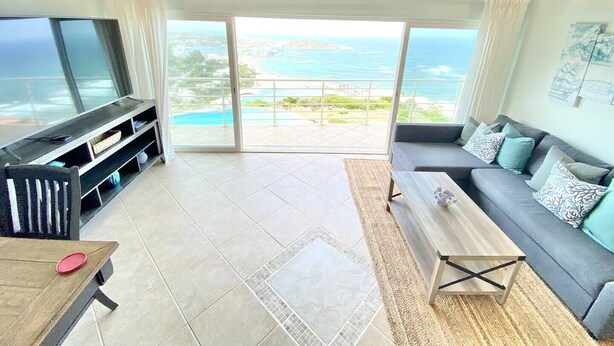 A spacious living room with a large TV, a sofa, and a coffee table. There is a sliding glass door leading to a balcony with a view of the ocean. - Luna Sul Mare - 2 Bed 2.5 Bath Condo with gorgeous ocean view! (Sint Maarten)