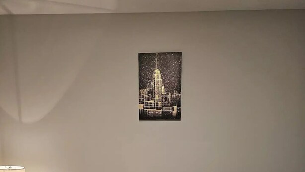Een hotelkamer met een opgemaakt bed, een nachtkastje met een lamp, en een lijst met een stadsbeeld aan de muur.