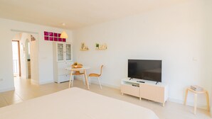 Une chambre d’hôtel moderne avec un grand lit, un téléviseur posé sur un meuble en bois, une table à manger avec des chaises et un coin cuisine visible à travers une arche.