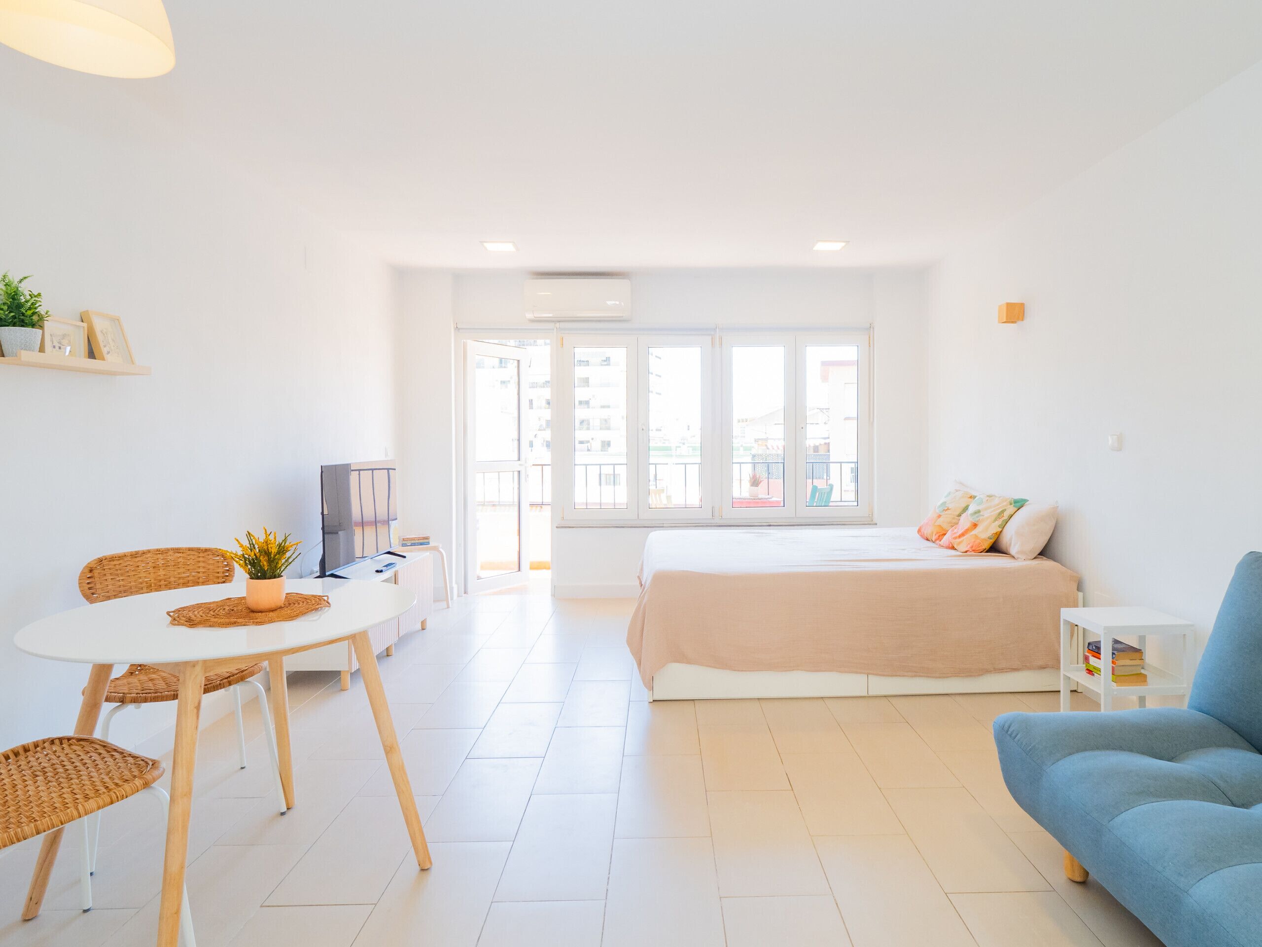 A bright, modern room with a bed, a blue sofa, a dining table with chairs, and a TV.