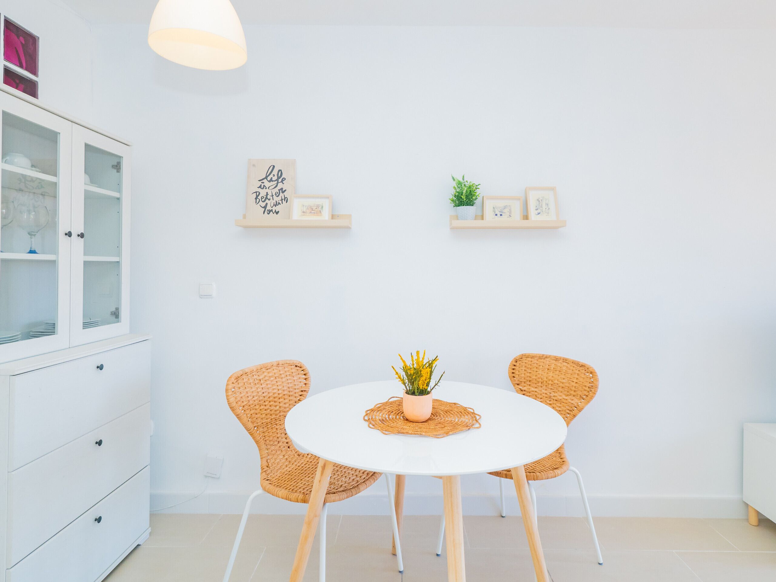 A modern dining area with a white table, two wicker chairs, a potted plant, and wall-mounted shelves with decorative items.