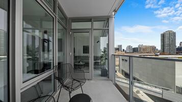 A modern balcony with glass doors, a seating area with chairs, and a view of the city skyline.