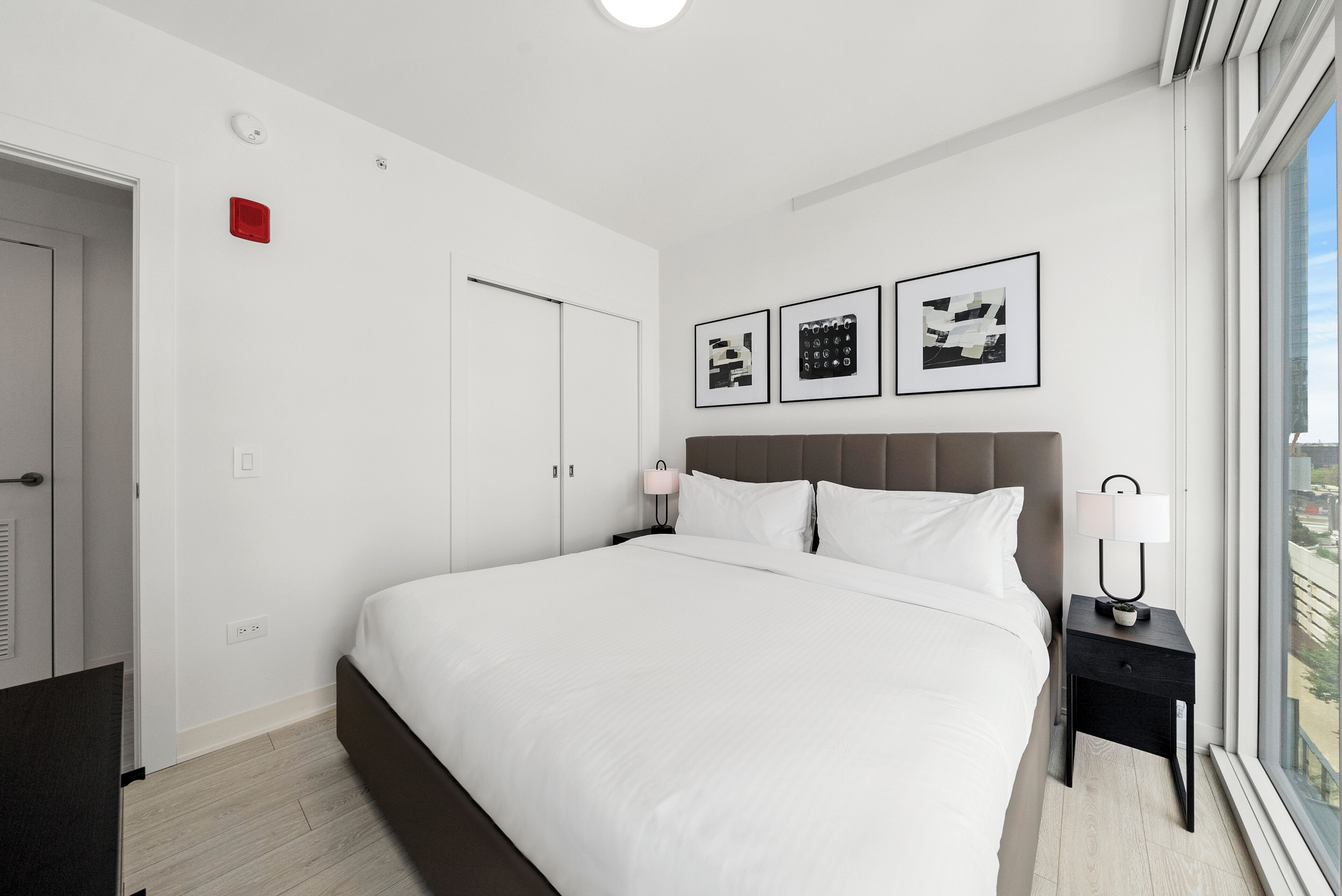 A modern hotel room with a large bed, a nightstand with a lamp, a window with a view, and a door.