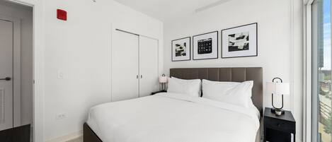 A modern hotel room with a large bed, a nightstand with a lamp, a window with a view, and a door.