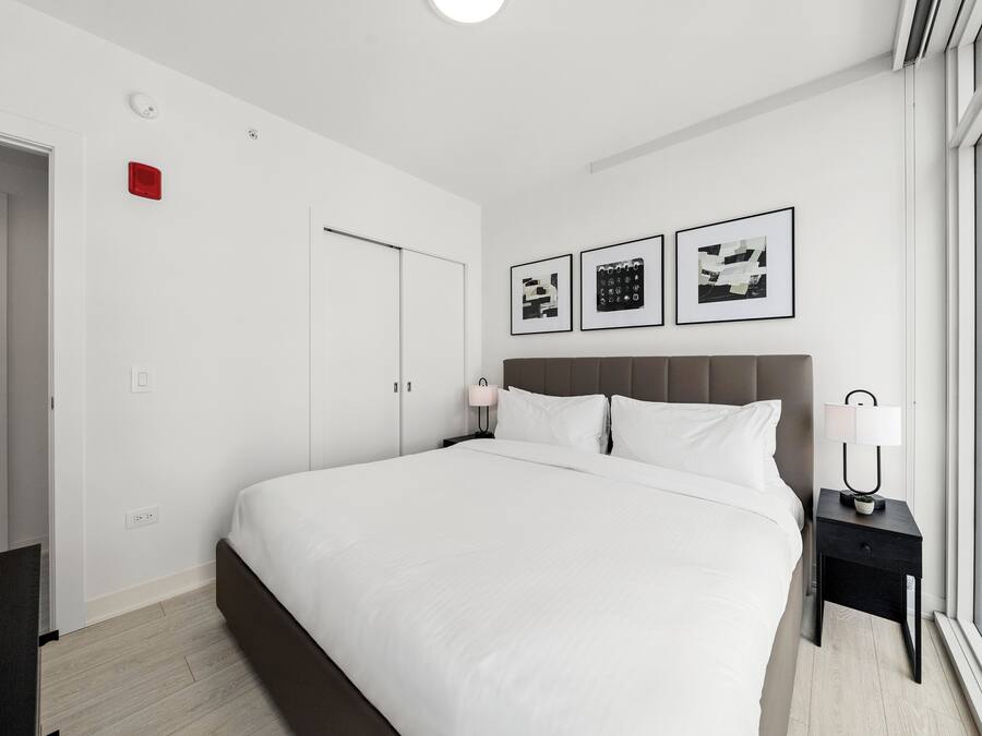 Una habitación de hotel moderna con una cama grande, una mesita de noche con lámpara, una ventana con vistas y una puerta.