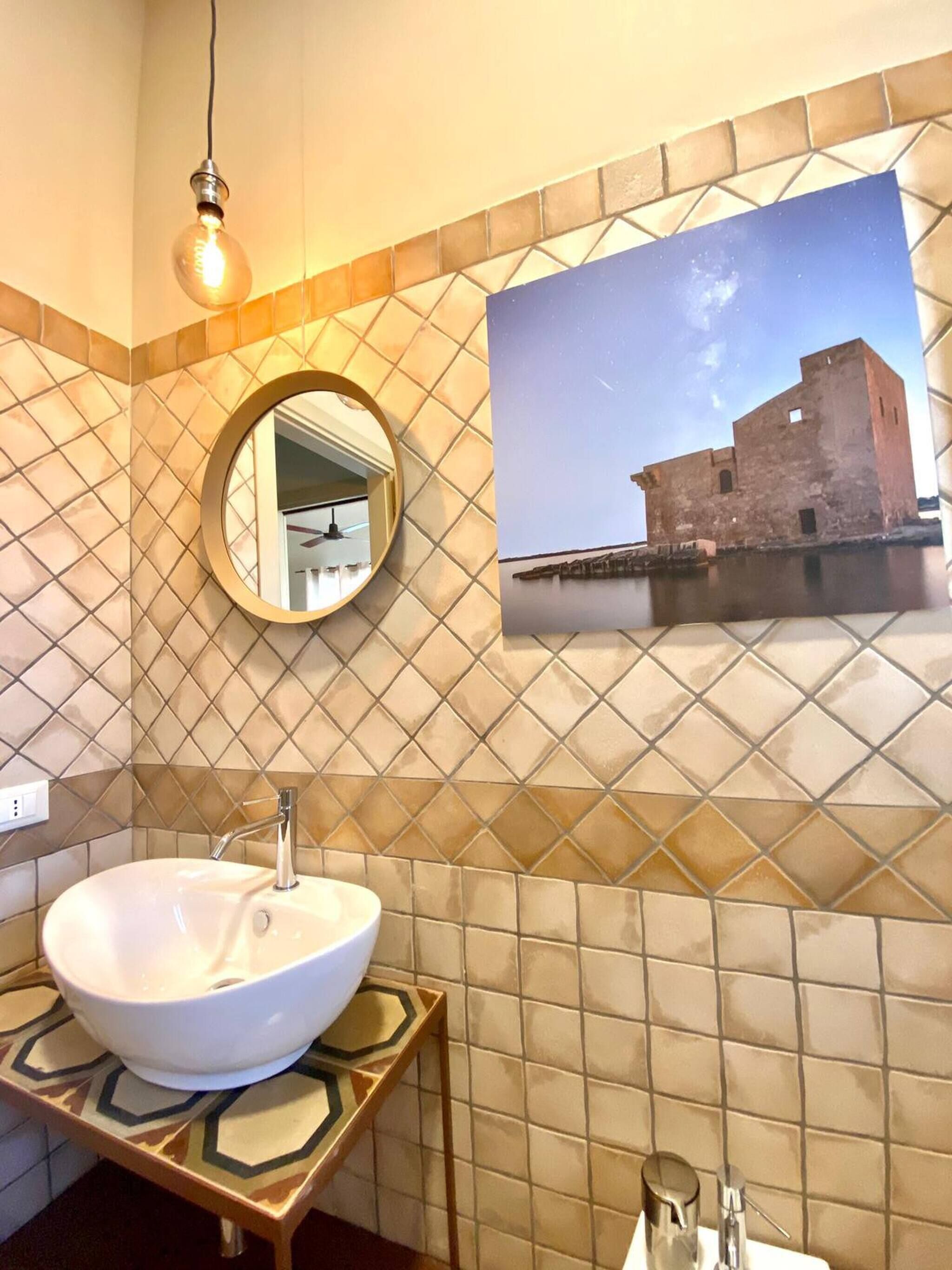 A bathroom with a large white sink, a round mirror, a wall-mounted picture of a historical building, and a hanging light bulb.
