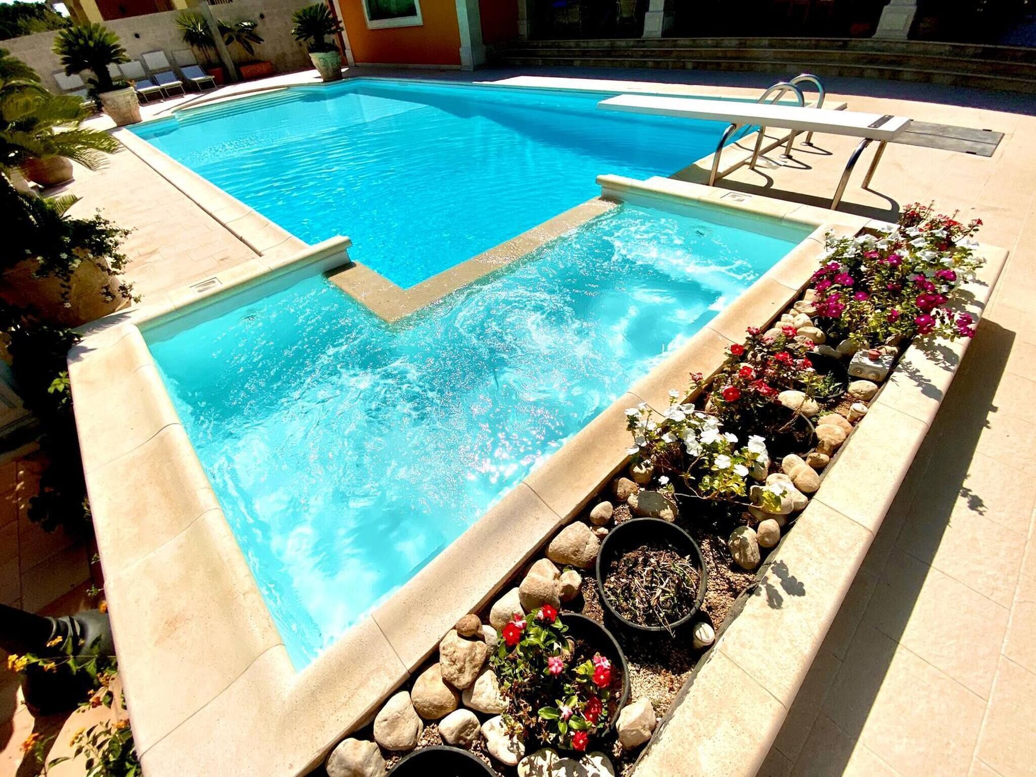 Ein rechteckiger Pool mit klarem, blauem Wasser, umgeben von einer gefliesten Terrasse und einem Blumenbeet mit verschiedenen Pflanzen und Blumen.