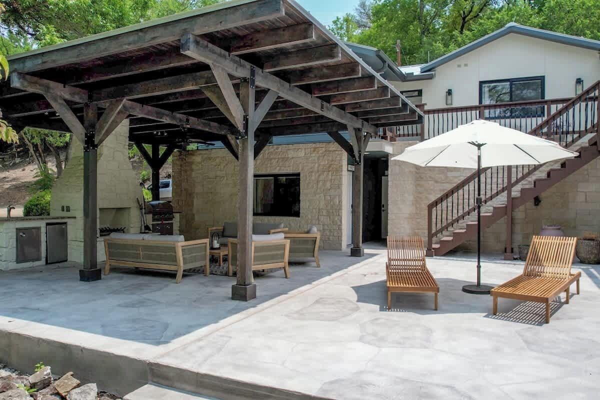 A covered outdoor patio with wooden furniture, a stone wall, and a staircase leading to a building.
