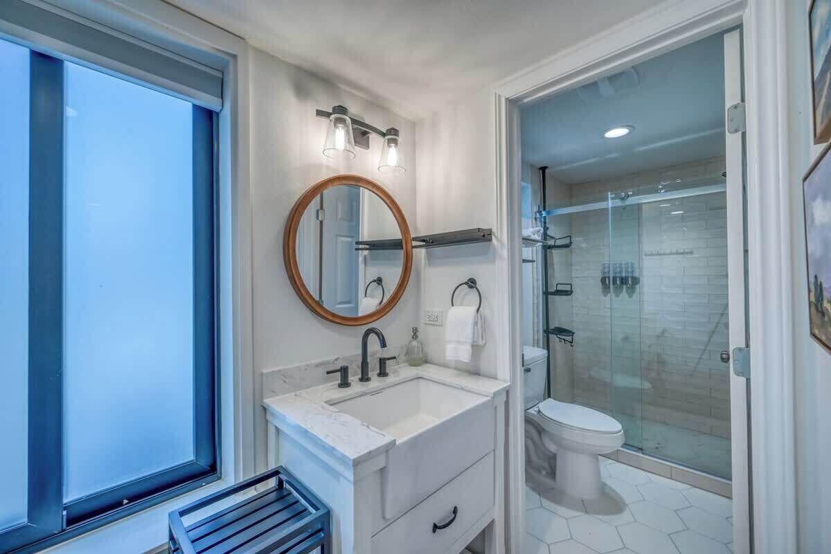 Une salle de bain moderne dotée d’une grande fenêtre, d’un miroir rond, d’un lavabo et d’une douche séparée par une paroi en verre.