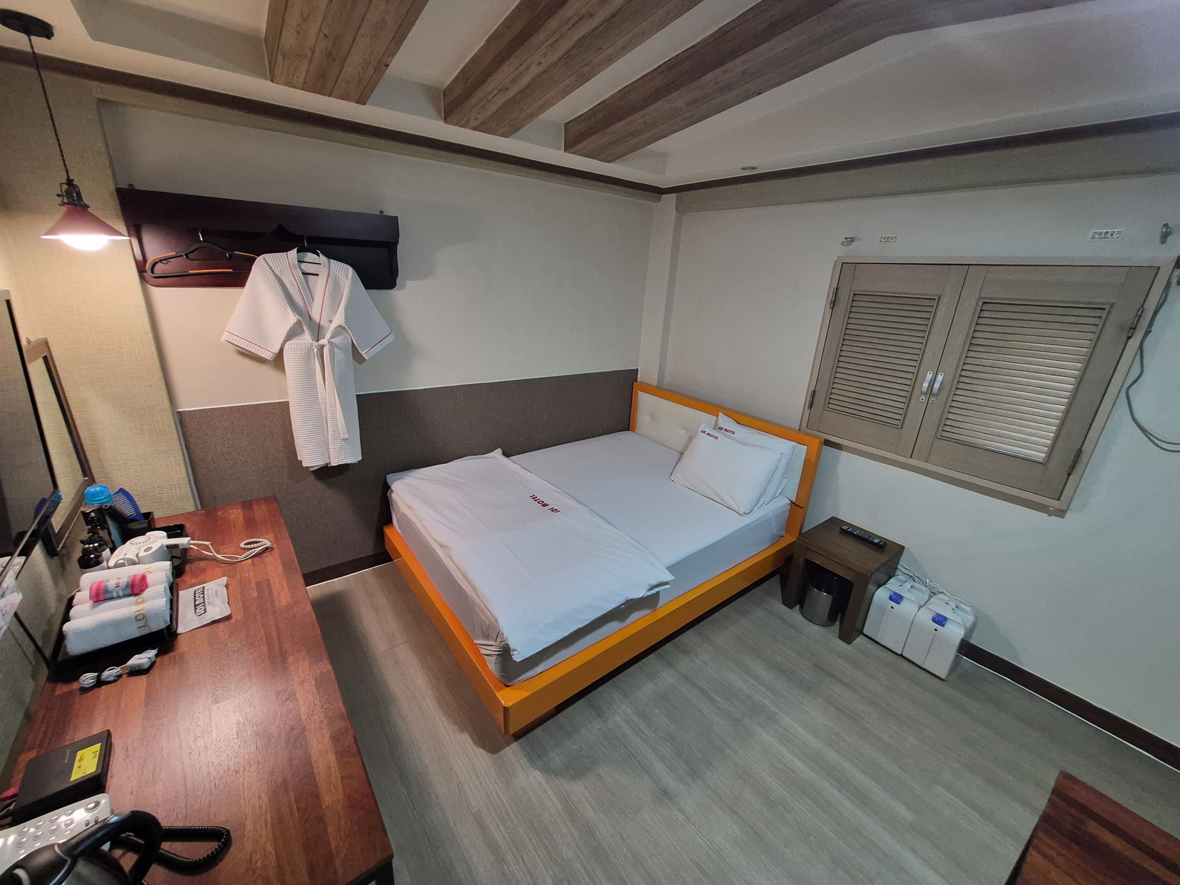 Una habitación de hotel con una cama de madera, una mesita de noche con teléfono, una bata colgada en la pared y una ventana con contraventanas.