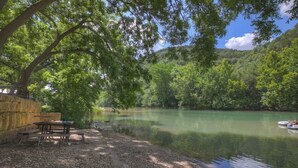 A riverside picnic area with tables and benches, surrounded by trees and a calm river. - Let's Go Tubing! Awesome House-next to River! (New Braunfels)