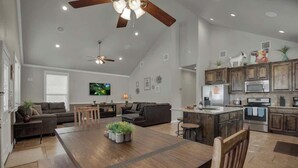 A spacious living room with a large wooden dining table, a ceiling fan, and a kitchen area with wooden cabinets and stainless steel appliances. - Let's Go Tubing! Awesome House-next to River! (New Braunfels)