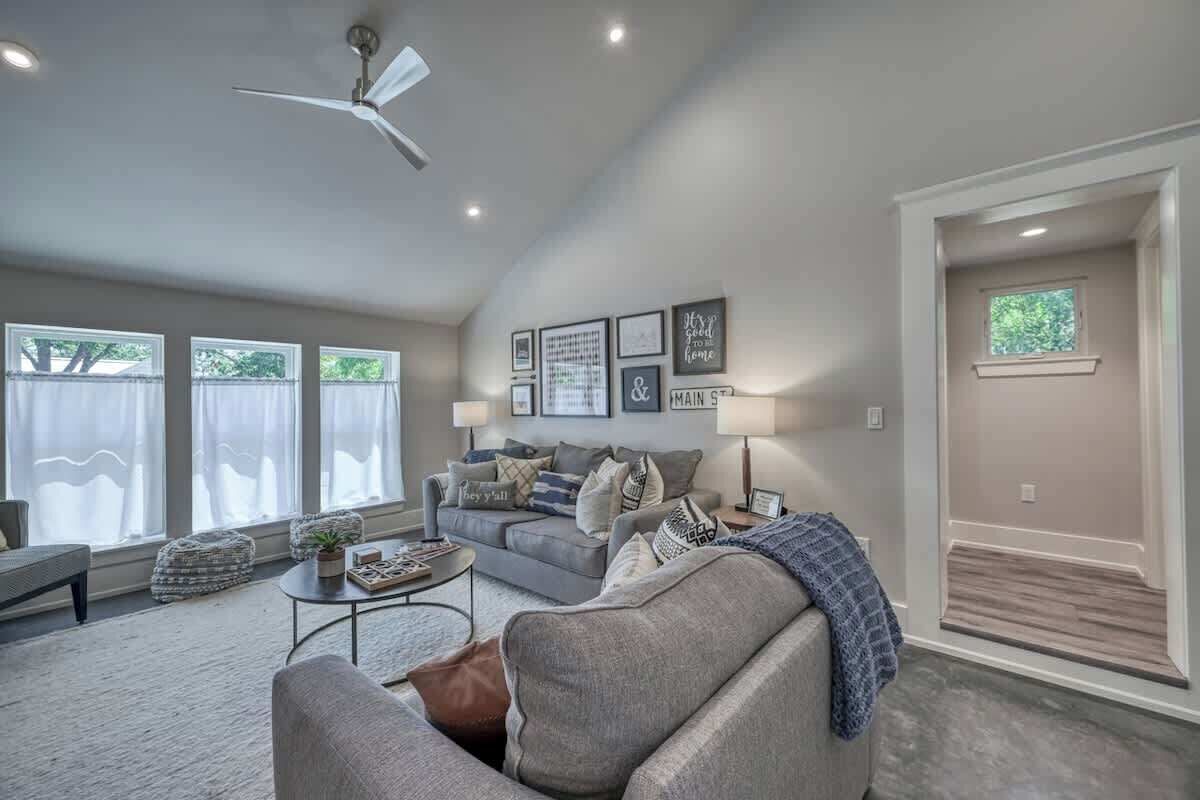 A spacious living room with a grey sofa, a coffee table, and a ceiling fan. There are framed pictures on the wall and a window with curtains.