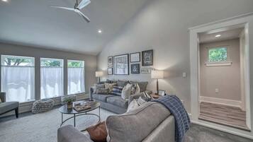 A spacious living room with a grey sofa, a coffee table, and a ceiling fan. There are framed pictures on the wall and a window with curtains.