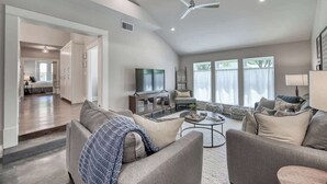 A spacious living room with a large sofa, a coffee table, and a television. There is a kitchen area visible in the background.