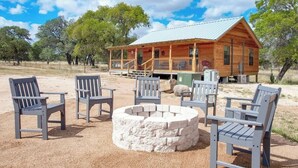 Une cabane en bois dotée d’un porche, entourée d’un espace extérieur aménagé avec des sièges et un foyer en pierre.