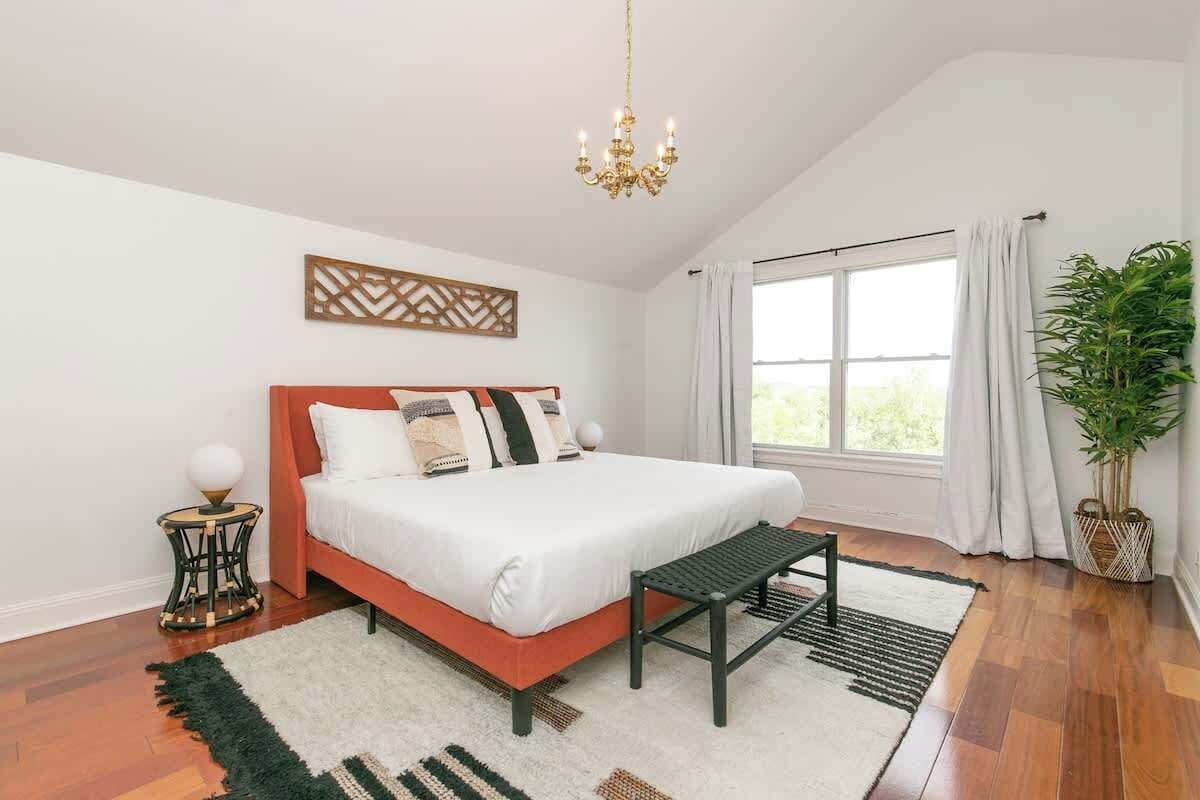 A bedroom with a bed, a bench, a chandelier, a window with curtains, a plant, and a decorative wall piece.