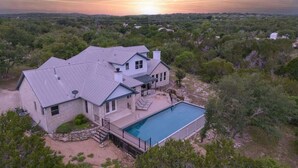 Une grande maison avec une piscine, entourée d’arbres et située dans un paysage vallonné.