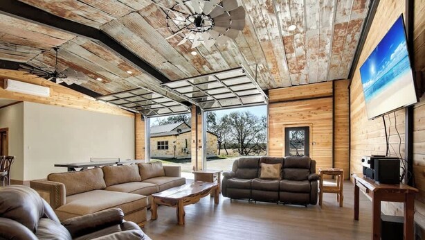 A spacious living area with a large sofa, a recliner, and a wooden coffee table. There is a television on a wooden stand and a ceiling fan. - Ranch Retreat Combo With Stunning Creek View-fire Pit (Fredericksburg)