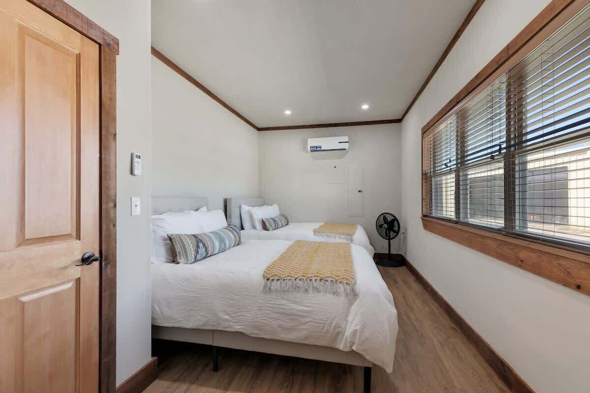 Une chambre avec deux lits, un plancher en bois, un ventilateur de plafond et une fenêtre avec des stores.